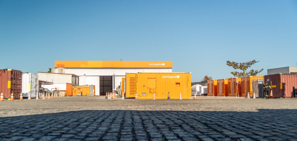 Um pátio industrial com geradores da Tecnogera dispostos sobre pisos de paralelepípedos. Cones de trânsito estão espalhados pelo local, destacando o fornecimento de energia feito pela Tecnogera, com um prédio estampado com o logotipo da empresa sob um céu azul e claro.