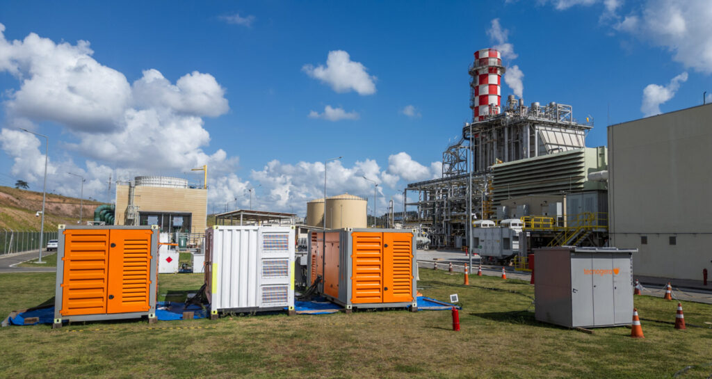 Três geradores laranja e um branco da Tecnogera sobre a grama, acompanhados de cones de trânsito e extintores de incêndio.