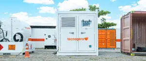 Gerador branco da Tecnogera ao ar livre, entre um tanque branco à esquerda e contêineres laranja e rosa à direita, sob céu azul.