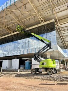 O Equipamento Tecnogera eleva dois profissionais até o teto de obra moderna, com painéis de vidro; materiais ao redor no chão.