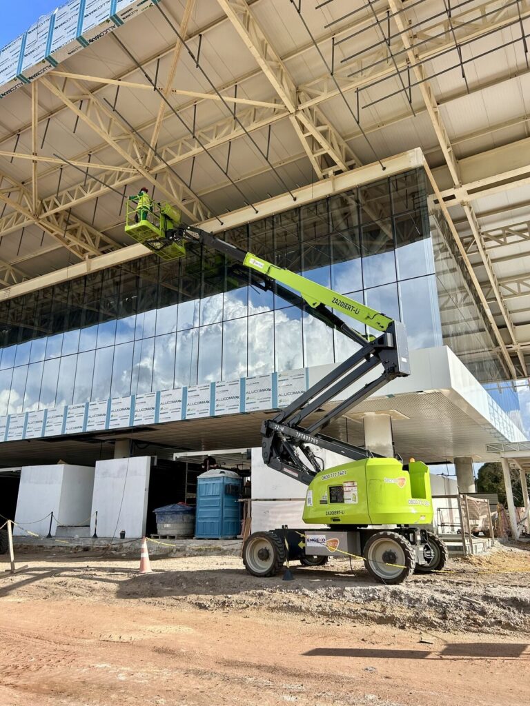 O Equipamento Tecnogera eleva dois profissionais até o teto de obra moderna, com painéis de vidro; materiais ao redor no chão.