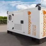 Gerador branco da Tecnogera ao ar livre, com telefone e localização rápida; vegetação e céu azul parcialmente nublado ao fundo.