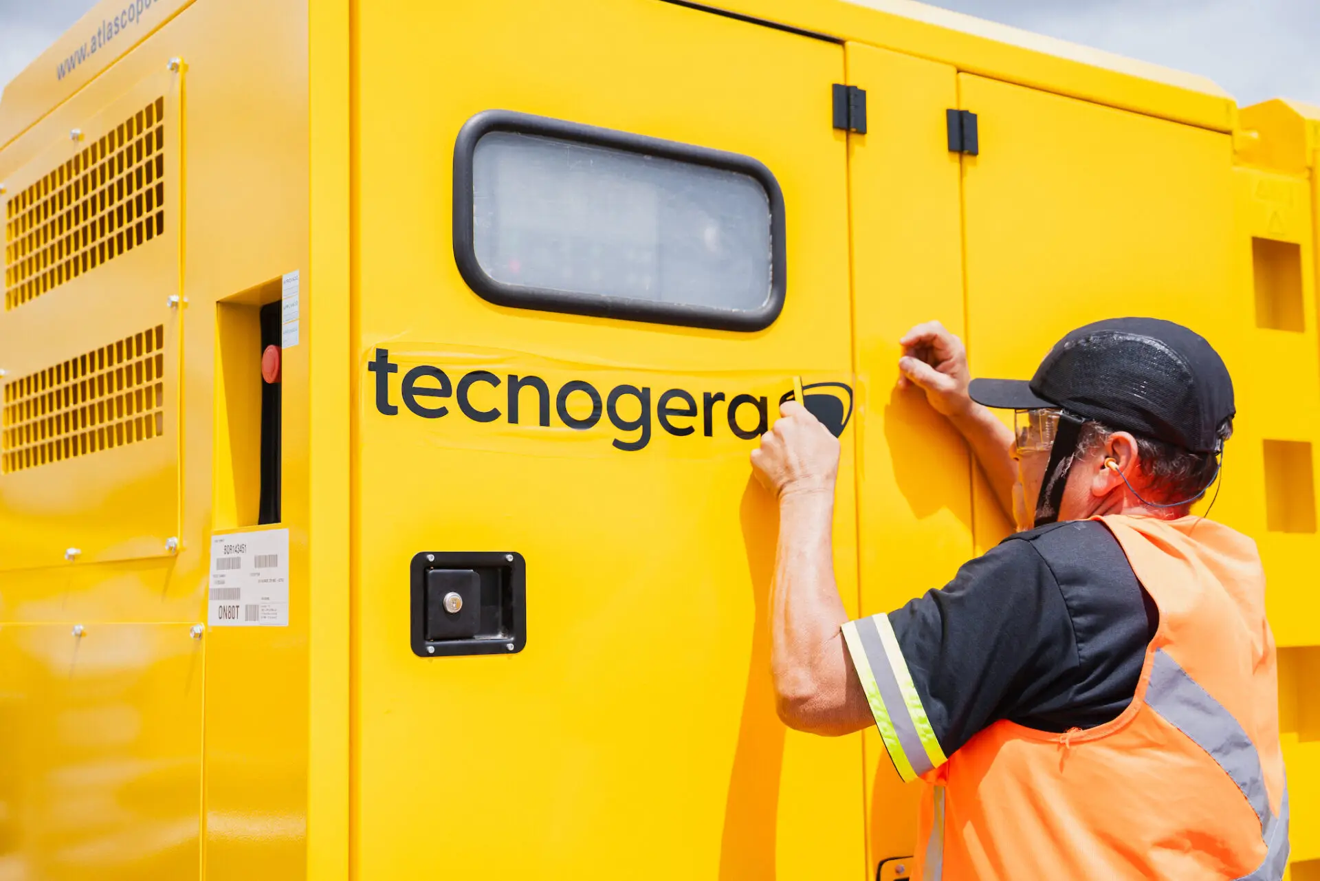 Colaborador com colete laranja e boné preto aplica adesivo Tecnogera em gerador amarelo sob sol, em ambiente externo.
