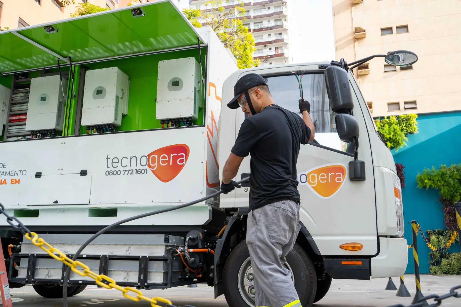 Um colaborador conecta cabos a um caminhão branco Tecnogera com painéis abertos, na rua, próximos a prédios e árvores.