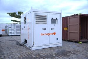 Banco de carga branco da Tecnogera ao ar livre, cercado por cabos, contêineres e bancos de carga; céu nublado ao fundo.