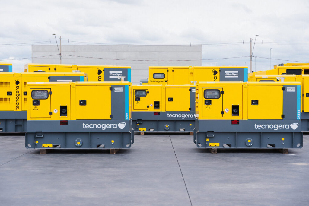Grandes geradores amarelos Tecnogera ao ar livre, ideais para data centers e garantindo energia contínua, com prédio ao fundo.