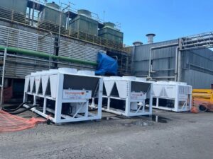 Três chillers industriais Tecnogera em área externa, com estruturas verdes ao fundo, tanques e tubulações junto a uma lona azul.