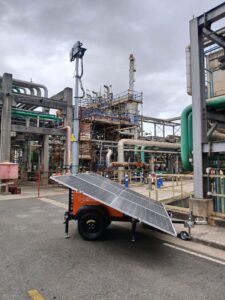 Unidade solar Tecnogera com antena e painéis ilumina obra noturna entre tubos e andaimes, sob céu nublado, com barreiras visíveis.